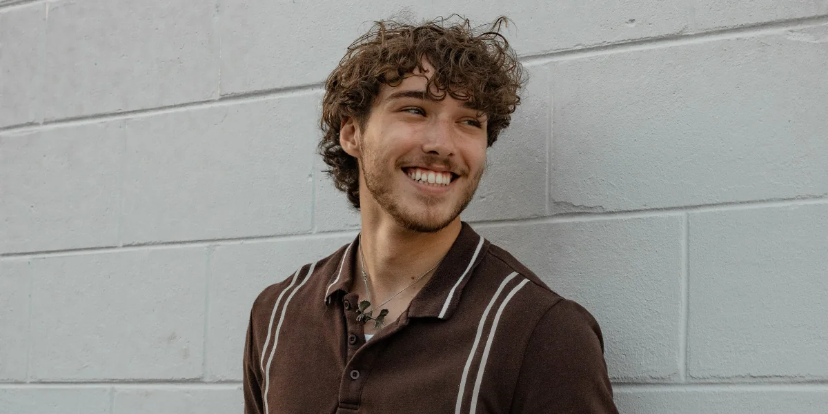 Curly-haired man smiling and looking to the side while resting against a wall