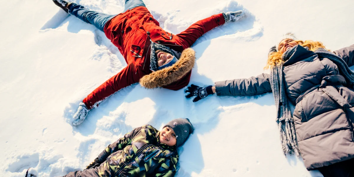three people one in red jacket while the other two in black thermal jackets laying down playing with snow