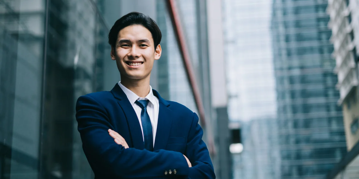 Smiling asian man in coat and tie with skyscraper behind him as a background.