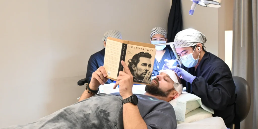 An international patient relaxes and reads a book while undergoing a DHI hair transplant procedure at Clinique de Paris, demonstrating the comfort and minimally invasive nature of the treatment.
